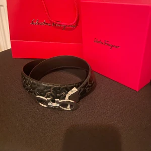Svart skärp från Salvatore Ferragamo - Säljer ett svart skärp från Salvatore Ferragamo med snyggt präglat mönster i lädret och en ikonisk silverfärgad dubbel-Gancini-spänne. Skärpet är stilrent och exklusivt, perfekt för att lyfta vilken outfit som helst. Kommer med originalförpackning och dustbag.