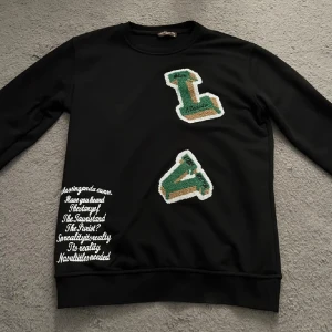 Louis Vuitton sweatshirt  - Svart sweatshirt med långa ärmar och coola broderade bokstäver i grönt, vitt och gult framtill. Tryckt vit text på ena sidan nedtill. Klassisk rund halsringning och mjukt material, perfekt för en avslappnad stil.
