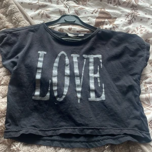 Svart croppad topp LOVE Lindex - Svart croppad topp från Lindex med stort silvrigt LOVE-tryck framtill. T-shirten har korta ärmar och en loose passform, perfekt för en chill och avslappnad stil. Tillverkad i mjuk bomull som känns skön mot huden.