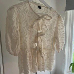 Krämvit blus med knyt från Gina Tricot - Säljer en krämvit blus från Gina Tricot i storlek M. Blusen har puffiga korta ärmar, strukturmönster i tyget och tre snygga knyt framtill. Den är lätt och luftig, perfekt för varma dagar och har en feminin, trendig look.