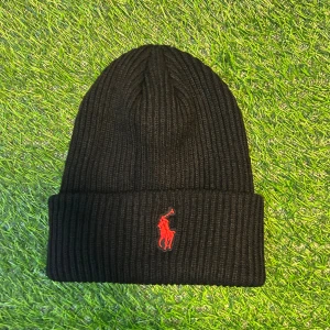 Svart ribbad mössa från Polo Ralph Lauren - Svart ribbstickad mössa från Polo Ralph Lauren med klassisk uppvikt kant och röd broderad logga framtill. Snygg och enkel design som funkar till alla jackor. Materialet är mjukt och värmande, perfekt för kyliga dagar.