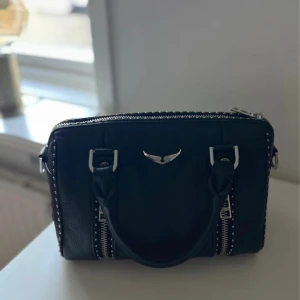 Svart handväska med nitar och dragkedja - Cool svart handväska från Rebecca Minkoff i skinn med silverfärgade detaljer, nitar längs kanterna och dubbla dragkedjor framtill. Väskan har två rejäla handtag och ett stilrent emblem i metall på framsidan. Perfekt storlek för det viktigaste.