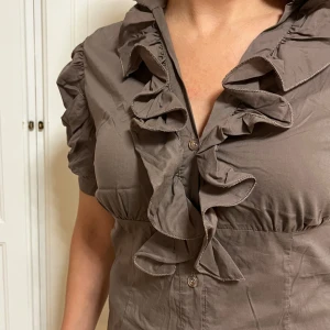 Brun volangskjorta från Laeticia Dreams - Supersnygg brun skjorta med volanger längs knäppningen och puffiga korta ärmar. Skjortan har en feminin passform och coola detaljer som ger extra edge. Perfekt för dig som gillar statement-plagg och vill sticka ut lite extra. Materialet känns lätt och bekvämt.