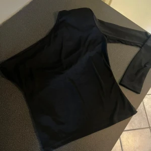 Svart oneshoulder topp - Säljer en svart oneshoulder topp med lång ärm på ena sidan. Snygg och stilren design som ger en cool asymmetrisk look. Materialet är mjukt och stretchigt, perfekt för att skapa en trendig outfit. Passar dig som gillar att sticka ut med enkla detaljer.