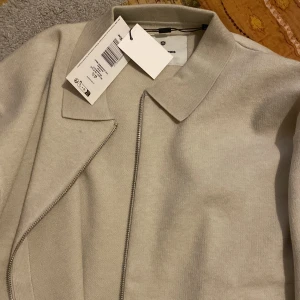 Beige cardigan jacka från Jack & Jones - Snygg beige cardigan jacka från Jack & Jones med stilren krage och diagonal dragkedja framtill. Jackan är långärmad och har en clean look som passar perfekt till din streetstyle. Materialet är mjukt och känns skönt mot huden.