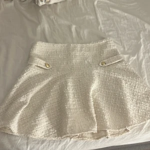 Vit kortkjol från H&M i tweed - Supersnygg vit kortkjol från H&M i tweed-look med två dekorativa fickor framtill och guldfärgade knappar. Kjolen är gjord av 100% återvunnen polyester och har dragkedja bak.