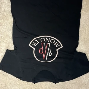 Svart Moncler t-shirt med stor logga - Svart t-shirt från Moncler med en stor, vit och röd logga tryckt på bröstet. Klassisk rund hals och korta ärmar. T-shirten har en clean look och är gjord i mjuk bomull som känns skön mot huden. Perfekt för dig som gillar exklusiva streetwear-vibbar.