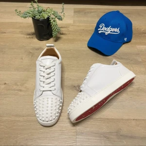 Christian Louboutin vita sneakers med nitar - Säljer ett par sjukt snygga vita sneakers från Christian Louboutin med coola nitar på tån och klassisk röd sula. Skorna har snörning och är tillverkade i slätt skinn. Perfekt för dig som vill sticka ut med en lyxig och edgy stil.