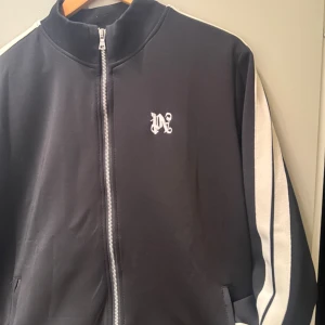 Svart sweatshirt zip från Palm Angels - Svart sweatshirt från Palm Angels med vita ränder längs ärmarna och broderad logga på bröstet. Jackan har silverfärgade dragkedjor både framtill och vid fickorna. Materialet är mjukt och känns som polyester. Perfekt för dig som gillar streetwear och snygga detaljer.