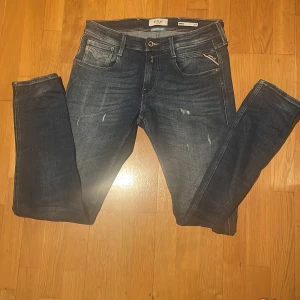 Replay Anbass  - Snygga Replay Anbass Slim Fit jeans i mörkblå tvätt med slitningar. Jeansen har en smal passform, normal midja och detaljer som Replay-logga på fickan och läderpatch bak. Perfekta för dig som gillar stilrena och moderna jeans. Hör av dig vid minsta fundering!!!