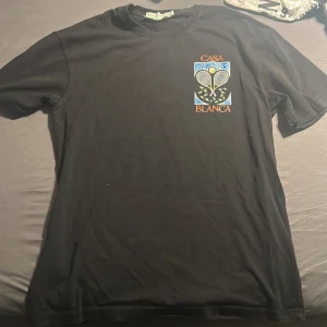 Svart Casablanca Tennis Club t-shirt - Svart t-shirt från Casablanca med stort tryck av två tennisracketar och texten 'Casa Blanca' på både bröstet och ryggen. Klassisk passform och rund hals. Perfekt för dig som gillar sportig stil och snygga prints.