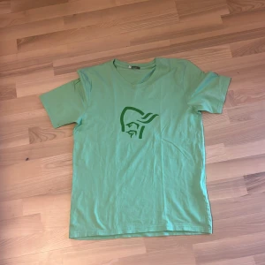 Grön t-shirt från Norrøna i bomull - Snygg grön t-shirt från Norrøna i storlek M. T-shirten har ett stilrent tryck med Norrønas logga på bröstet och en mindre logga i nacken. Klassisk passform med rund hals och korta ärmar. Perfekt för en avslappnad look.