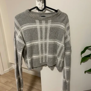 Grå croppad rutig stickad tröja - Säljer en grå stickad tröja med vitt rutmönster. Tröjan är croppad med lång ärm och har en ribbad rund halsringning. Perfekt för dig som gillar en chill och trendig stil.