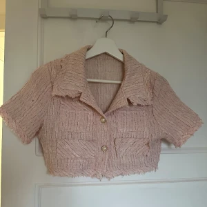 Croppad rosa bolerotopp med pärlknappar - Säljer en croppad bolerotopp i ljusrosa med korta ärmar och krage. Toppen har två fickor framtill och pärlknappar som detalj. Materialet är grovvävt och ger en lyxig känsla, med fransiga kanter för en cool touch. Perfekt att styla med matchande kjol. Matchade sett