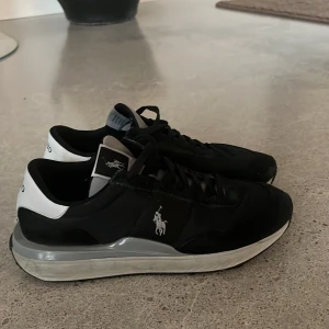 Svarta sneakers från Polo Ralph Lauren - Svarta sneakers från Polo Ralph Lauren med vit logga på sidan och vit häl. Skorna har snörning, platt sula och en sportig design. Ovandelen är i syntet och skinn, med detaljer i grått och vitt. Perfekta för en stilren och avslappnad look.