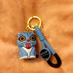 Blå KPOP nyckelring med katt - Nyckelring med en söt blå kattfigur med stora gula ögon och ett hjärta på magen. Nyckelringen har guldfärgad ring och blå flätad detalj med svart och blå KPOP Girl Group-märke. Perfekt accessoar för KPOP-fans.