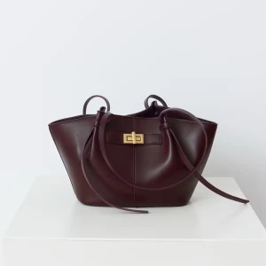 Mörkbrun axelväska i skinn med gulddetalj - Snygg mörkbrun axelväska i skinn med stilren, strukturerad form och långa remmar. Väskan har en elegant guldfärgad vridlåsdetalj framtill och minimalistisk design som ger en lyxig vibe. Perfekt storlek för att få plats med allt du behöver.