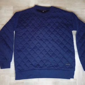 Marinblå quiltad tröja från Smog - Quiltad marinblå tröja från Smog i storlek XS. Tröjan har ett snyggt rutmönster över hela ytan, rund halsringning och långa ärmar med ribbade muddar. Perfekt för dig som gillar stilrena och bekväma plagg med lite extra struktur.