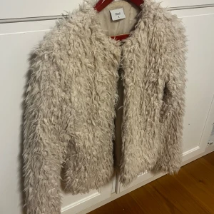 Beige pälsjacka från Grunt - Fluffig och trendig beige pälsjacka från Grunt i storlek barn XL alltså S i vanlig storlek Jackan har långärmade ärmar och en cool, oversized look med mjukt foder på insidan. Perfekt för dig som vill sticka ut med en statement piece i garderoben.