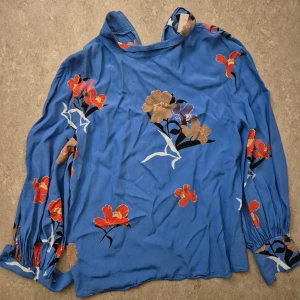 Blus - Snygg blå blus från H&M med blommigt mönster i rött, beige och lila. Blusen har långa ärmar med resår vid handlederna och en dold dragkedja i ryggen. Perfekt för dig som vill ha en färgstark och trendig look.