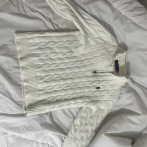 Vit kabelstickad tröja Polo Ralph Lauren - Säljer en vit kabelstickad tröja från Polo Ralph Lauren med half zip och långärmad design. Tröjan har ribbade muddar och krage, samt den klassiska broderade loggan på bröstet. Materialet är mjuk bomull, perfekt för kyliga dagar.