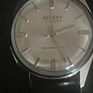 Vintage Regent silverfärgad klocka med svart band - Stilren klocka från Regent med silverfärgad urtavla och markerade index. Boetten är rund och i metall, medan armbandet är svart och har en klassisk look. Perfekt för dig som gillar enkel och tidlös design.