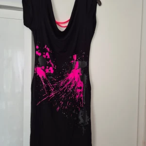 Klänning - Svart kortklänning med coolt neonrosa och grått stänkmönster framtill. Klänningen har korta ärmar, rundad hals med drapering och snygga rosa band i ryggen. Perfekt för dig som vill sticka ut med en edgy och färgstark look.