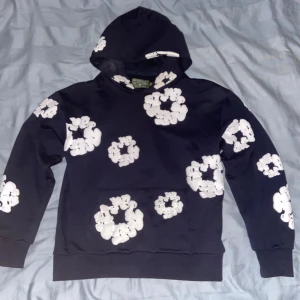 Mörk blå hoodie med vita blommor Denim Tears - Mörk blå hoodie från Denim Tears med stora vita blommor tryckta över hela tröjan, inklusive huvan. Tröjan har huva med snörning och en klassisk känguruficka framtill. Mjuk bomullskvalitet och långärmad passform som ger en chill vibe.