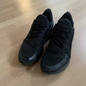 Svarta Nike Air Max 270 sneakers - Säljer ett par helsvarta Nike Air Max 270 sneakers med mesh-överdel och synlig Air-enhet i hälen. Skorna har snörning, rund tå och en sportig vibe. Perfekta för dig som gillar streetstyle och vill ha bekväma skor med snygg design.