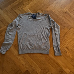 Grå v-ringad tröja Lexington L - Grå v-ringad tröja från Lexington i storlek L. Tröjan har lång ärm och ribbade muddar vid ärmslut och nederkant. Mjuk och skön stickad känsla, perfekt för chill dagar. Diskret Lexington-logga på bröstet.