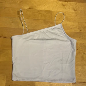 Vit oneshoulder topp från NA-KD - Säljer en stilren vit topp från NA-KD i oneshoulder-modell med tunna axelband. Toppen är croppad och har en clean look som är perfekt för att skapa en trendig outfit. Materialet är mjukt och stretchigt, vilket gör den bekväm att bära. Taggen sitter kvar.