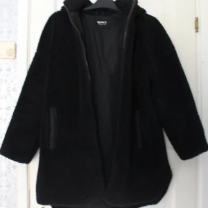 Svart teddyjacka från ONLY  - Mysig svart teddyjacka från ONLY i storlek XS. Jackan har huva, dragkedja framtill och praktiska fickor. Materialet är fluffigt och värmande polyester, perfekt för kyliga dagar. Enkel och stilren design som passar till många olika outfits.