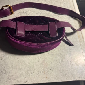 Lila quiltad crossover axelväska i sammet - Säljer en rund lila axelväska med quiltad material i mjuk sammet och matchande lila axelrem i skinn. Väskan har dragkedja i guld och ett ljust innerfoder. Perfekt storlek för det viktigaste och snygg detalj till din outfit. Perfekt för strand häng, city liv. Väldigt fint skick då jag har använt en annan. 