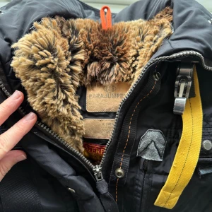 Svart Parajumpers jacka med pälsfoder - Svart jacka från Parajumpers med fluffigt brunt pälsfoder, gul detaljrem och flera coola fickor. Jackan har dragkedja, lång ärm och robusta metalldetaljer. Perfekt för dig som vill ha en snygg och funktionell vinterjacka med streetkänsla.
