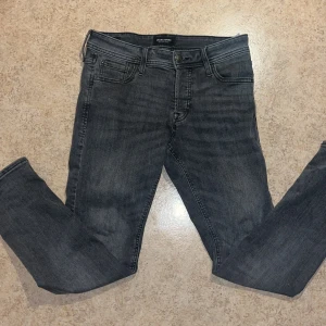 Grå slim jeans från Jack & Jones - Snygga grå jeans från Jack & Jones i modellen slim/Glenn. De har en slim passform med klassiska fem fickor och svarta detaljer. Jeansen är tillverkade i stretchigt denim för extra komfort och har en mörkgrå tvättad look.