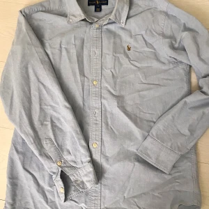 Ljusblå skjorta från Ralph Lauren - Snygg ljusblå skjorta från Ralph Lauren med klassisk krage och knappar framtill. Ikoniska broderade loggan på bröstet ger en stilren touch. Skjortan har långa ärmar och är gjord i mjuk bomull som känns skön mot huden. Perfekt för dig som gillar enkel och tidlös stil.