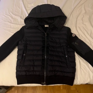 Svart Moncler pufferjacka med huva - Svart pufferjacka från Moncler med stickade ärmar och quiltad kropp. Jackan har huva, dragkedja framtill och Moncler-logga på ärmen. Fickor med tryckknappar och extra ficka med dragkedja på ena ärmen. Perfekt för kyliga dagar.den känns som en S i storlek 