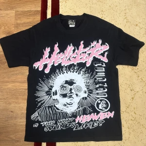 Hellstar svart graphic t-shirt - Svart t-shirt från Hellstar med stor graphic print framtill i vitt och rosa, samt Hellstar Records-tryck på ryggen. T-shirten har en klassisk passform med korta ärmar och rund hals. Materialet är mjuk bomull, perfekt för dig som gillar bold streetwear.