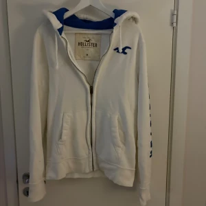 Vit hoodie från Hollister med blå detaljer - Vit hoodie från Hollister med blå logga på bröstet och blå text längs ena ärmen. Tröjan har dragkedja, huva med blå insida och två fickor framtill. Perfekt för chill dagar och snygg streetstyle. Riktigt snygg och ovanlig tröja!