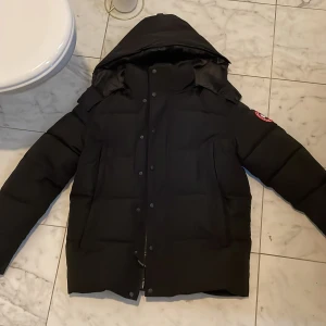 Svart dunjacka från Canada Goose - Säljer en svart dunjacka från Canada Goose med stor huva och snygg pälsdetalj. Jackan har klassisk patch på ärmen, dragkedja och knappar framtill samt flera fickor. Perfekt för kalla dagar och riktigt varm. Passformen är normal och jackan är långärmad.