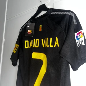 Barcelona David Villa #7 Nike fotbollströja - Säljer en svart FC Barcelona fotbollströja med David Villa och nummer 7 i gult på ryggen. Tröjan har officiella LFP och sponsorpatchar, samt klubbmärke och Nike-logga framtill. Materialet är lätt och andas, perfekt för match eller träning.