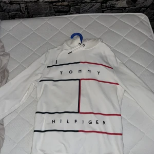 Vit hoodie Tommy Hilfiger S - Snygg vit hoodie från Tommy Hilfiger i ekologisk bomull. Tröjan har långa ärmar, huva med snörning och coolt grafiskt tryck i svart och rött på framsidan. Klassisk Hilfiger-logga på ärmen och stilren design som sticker ut.