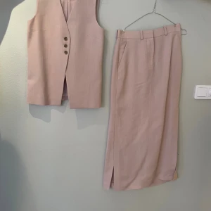 Beige lång väst med knappar - Stilren lång väst i beige med tre knappar framtill och diskreta fickor. Västen har en rak passform och är ärmlös, perfekt att styla över en skjorta eller t-shirt. Gjord i ett mjukt tyg som känns lyxigt och lätt.