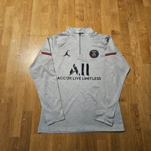 PSG långärmad träningströja Jordan - Snygg långärmad PSG träningströja i ljusgrått med svarta prickar, tillverkad i lätt och ventilerande material. Tröjan har halv dragkedja, PSG-logga på bröstet, Jordan-logga och tryckta detaljer som 'Accor Live Limitless' och 'Visit Rwanda'. Röd, vit och blå rand på ärmarna.