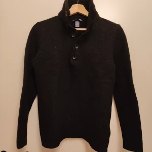 Svart stickad tröja med hög krage H&M - Svart stickad tröja från H&M med hög krage och fyra snygga knappar framtill. Tröjan har långärmade ärmar med förstärkta armbågslappar och ribbade muddar. Materialet är 80% ull och 20% polyamid, vilket gör den både mjuk och varm. Perfekt för dig som gillar stilren och enkel design.