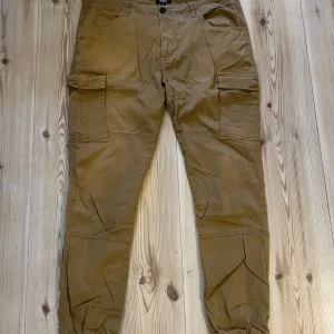 Beiga cargopants från William Baxter strl.L - Säljer ett par snygga beiga cargopants från William Baxter i storlek L. Byxorna har flera praktiska fickor på sidorna och bak, samt resår vid bensluten för en chill look. Perfekta för dig som gillar streetstyle och funktionella detaljer.