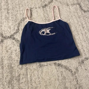 Mörkblå linne från MFPSHIF - Säljer ett mörkblått linne med tunna rosa axelband och en vit stjärnlogga framtill från MFPSHIF. Toppen har en croppad passform och är perfekt för dig som gillar sportig stil. Materialet känns lätt och skönt mot huden.