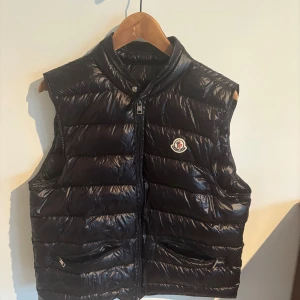 Svart dunväst från Moncler - Säljer en svart, glansig dunväst från Moncler med ståkrage och dragkedja framtill. Västen har två fickor med dragkedja och ett broderat Moncler-märke på bröstet. Modellen är quiltad och har en stilren look som passar till många outfits.