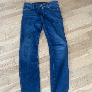 Blå Replay jeans, storlek 26/30 - Snygga blå jeans från Replay med klassisk femficksdesign och silverfärgad knapp. Jeansen har raka ben och normal passform, med tydliga kontrastsömmar och metallmärke vid fickan. Perfekt för dig som gillar stilrena och tidlösa jeans.