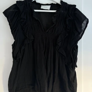 Svart volangblus från Neo Noir - Supersöt svart blus från Neo Noir med volangdetaljer längs ärmhålen och v-ringning. Perfekt att styla med jeans eller kjol. 